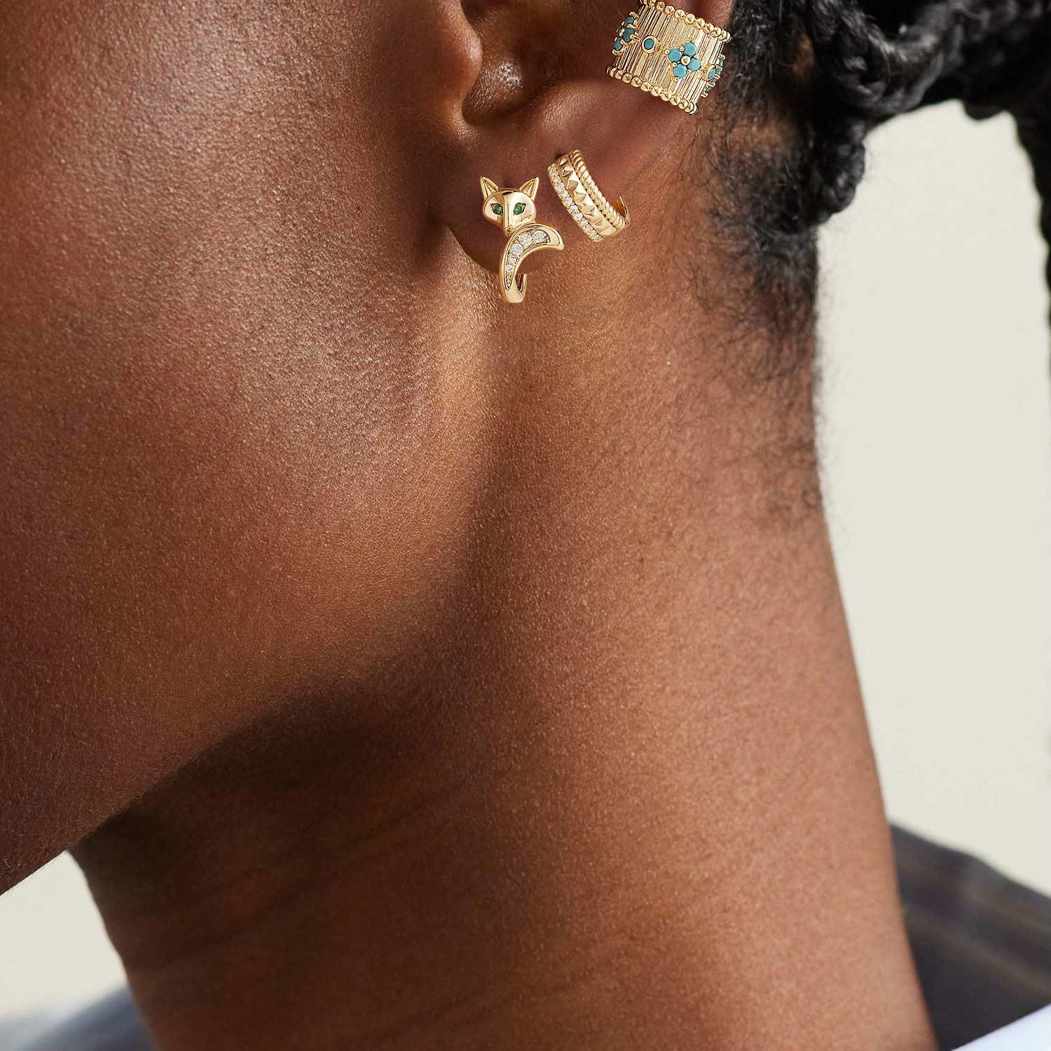 Side close-up of a woman wearing playful gold fox hoop earrings, showcasing the green eyes and dainty design on her ear.